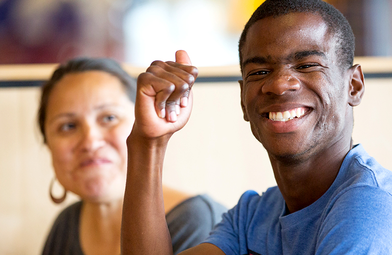 A young black man with a disability smiles at the camera. His a woman who provides him with direct care smiles at him. She has dark brown hair and medium skin.