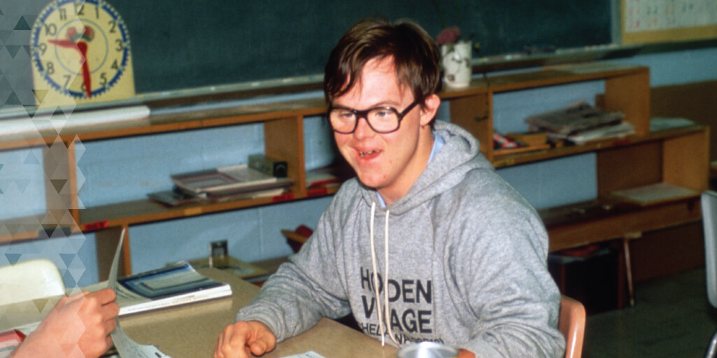 David smiles in a classroom. He is wearing glasses and a gray sweatshirt that says Holden Village.