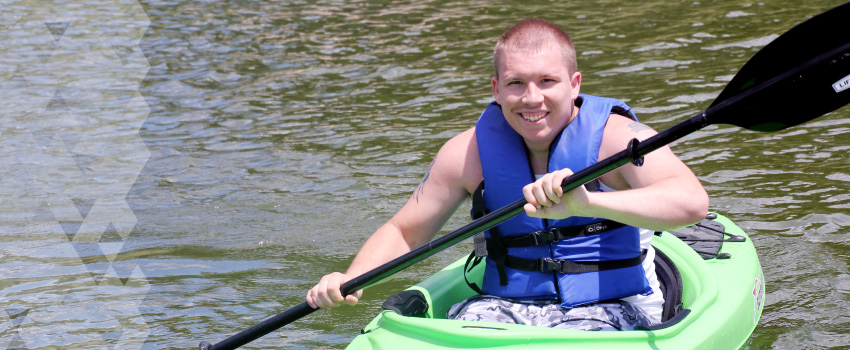 A young man paddles in a kayak on a lake. He is smiling.