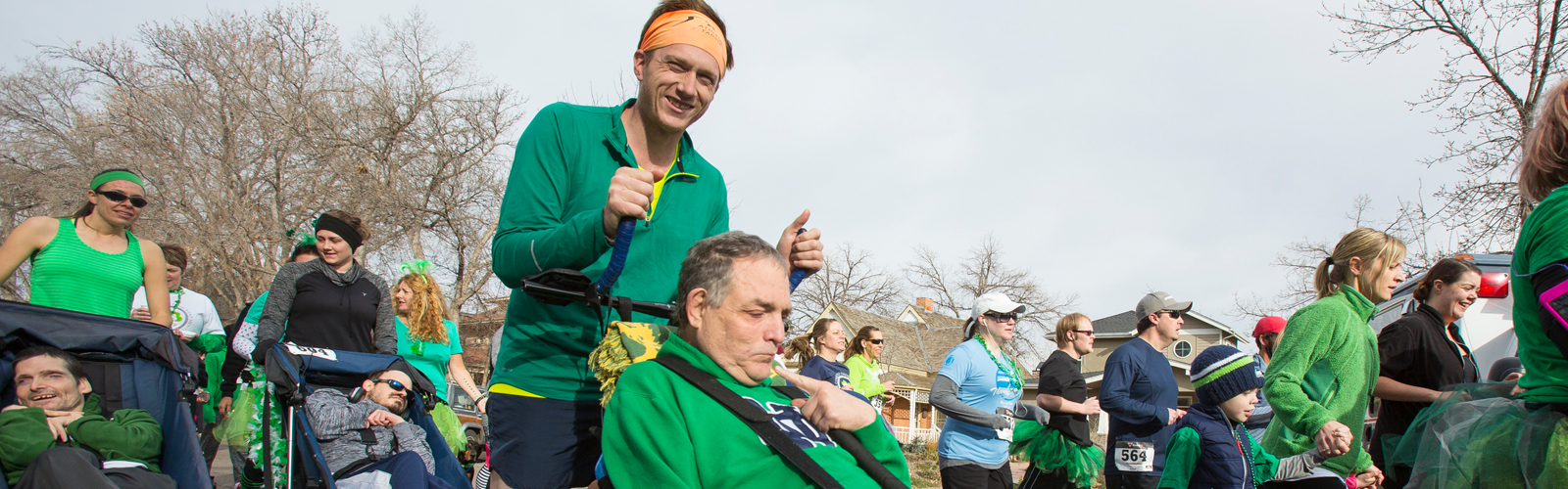 A young man wearing a green pullover and orange bandana on his head pushes an older man, also wearing a green sweatshirt, in a road race.