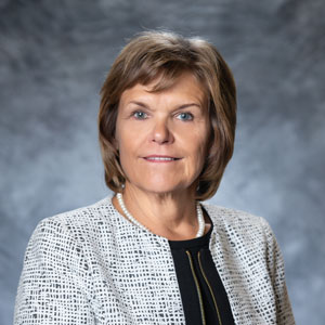 A woman with medium light skin and dark blond hair. She is wearing a black blouse and a gray suit jacket.