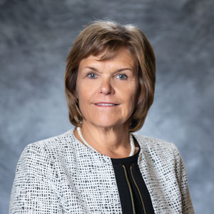 Pat-Nimtz-square A woman with medium light skin and dark blond hair. She is wearing a black blouse and a gray suit jacket.